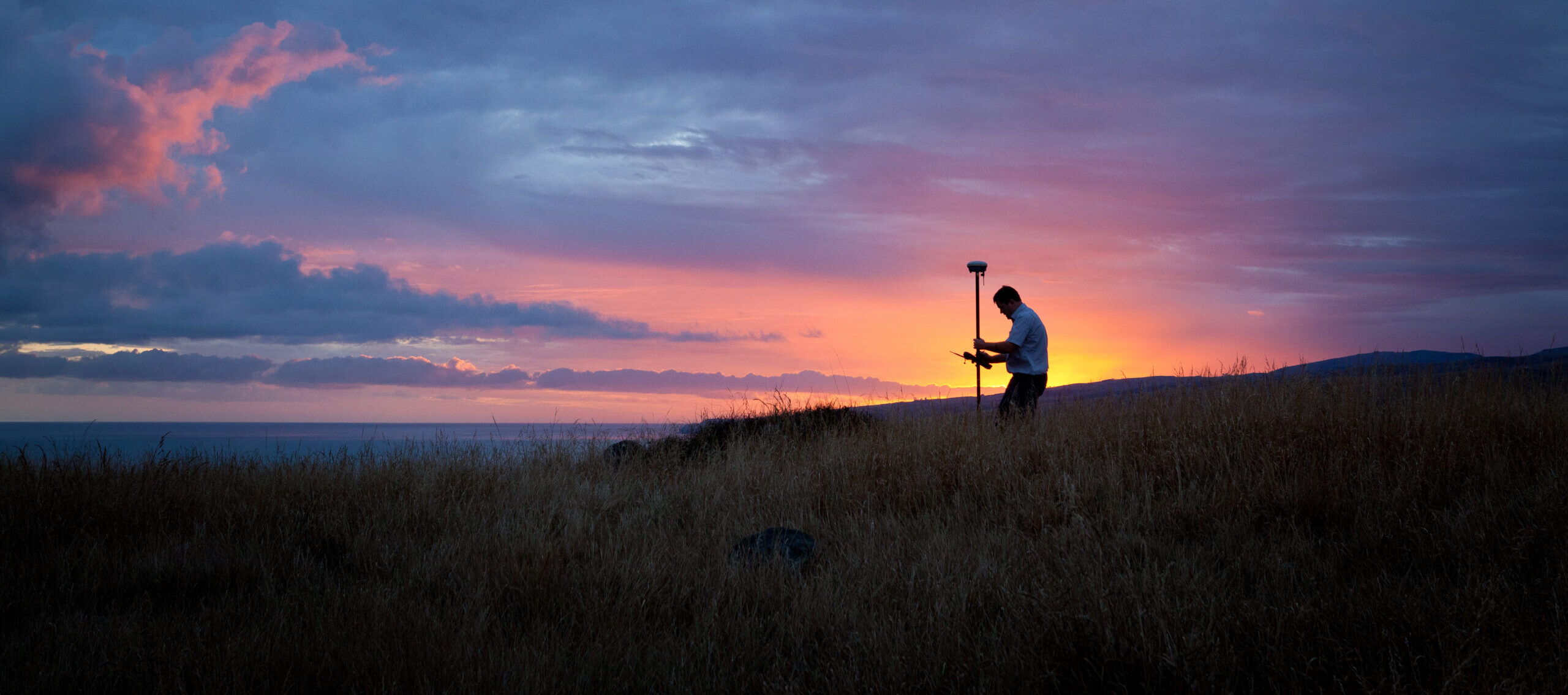 Person holding a GPS survey pole in a grassy field at sunset.